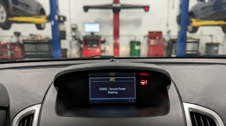 Chevrolet dashboard showing a power steering warning light and diagnostic trouble code with a blurred service garage in the background.